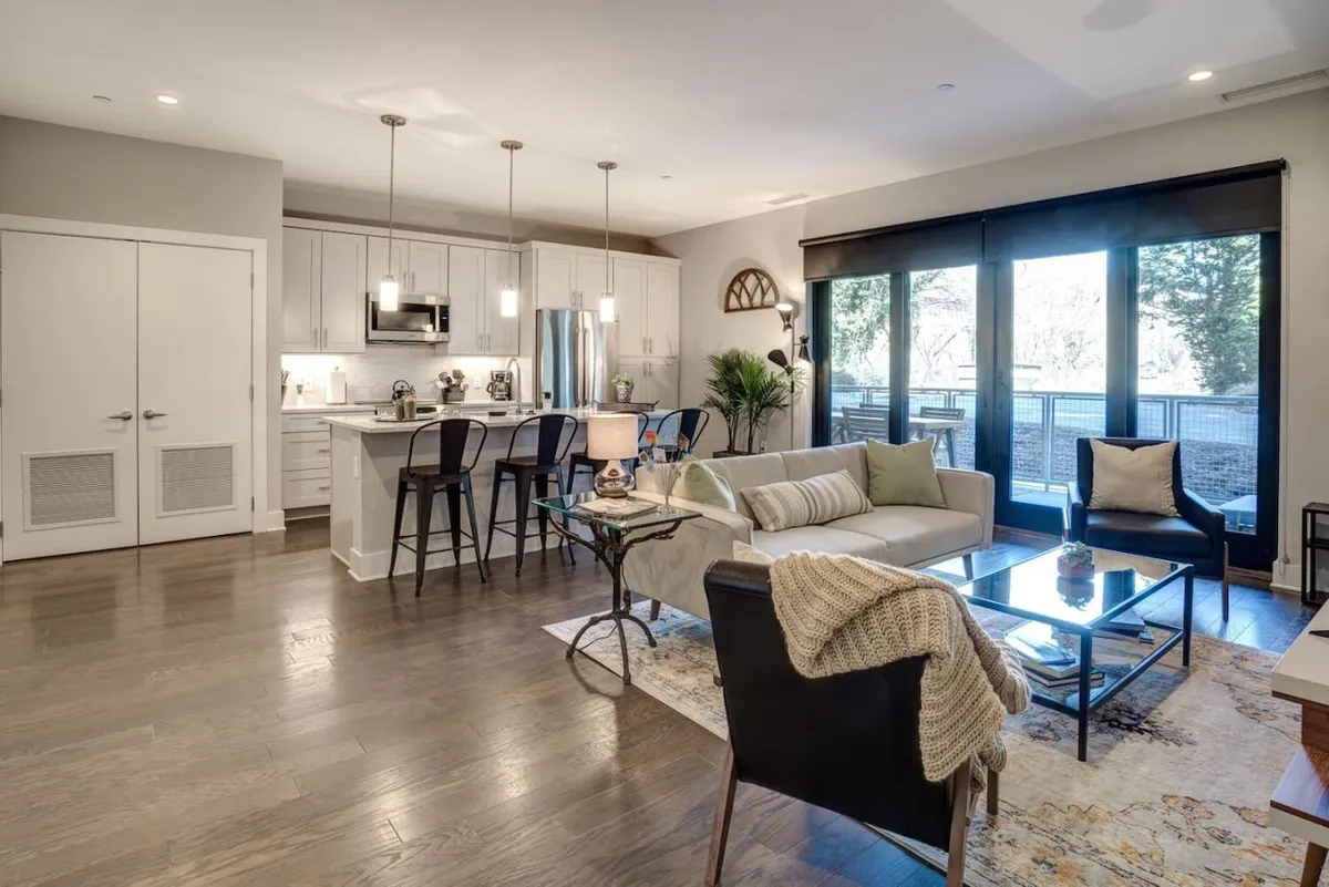 This image captures an open-concept living and dining area with a comfortable sofa, a kitchen island, a dining table, and expansive large windows that flood the space with natural light. The room is styled with contemporary decor and offers clear sightlines between living, dining, and kitchen spaces, creating a cohesive and welcoming atmosphere.