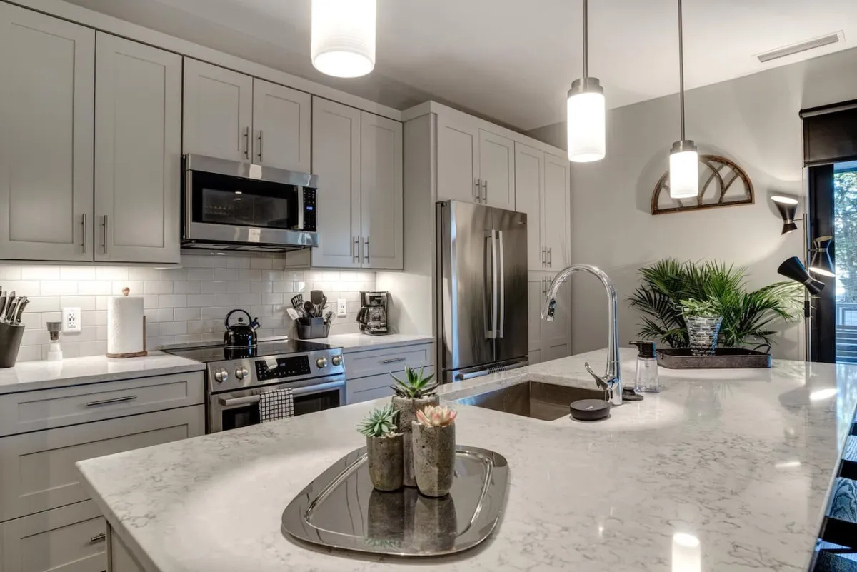 The scene features a kitchen island with a sleek countertop, pendant lighting overhead, gleaming stainless steel appliances, and carefully placed decorative plants. The image conveys a space ready for anything from preparing a family breakfast to hosting an intimate dinner, with a cozy and functional vibe.