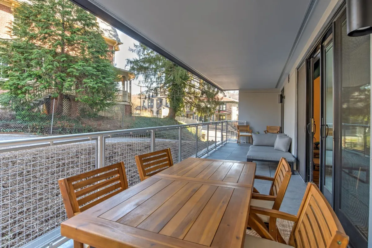 An inviting outdoor patio features a wooden dining table and a comfortable seating area, framed by lush greenery in the background. The space is bathed in warm light, suggesting a peaceful environment perfect for relaxing or entertaining. The view hints at the vibrant city life just beyond the greenery, creating a seamless transition between private retreat and urban adventure.