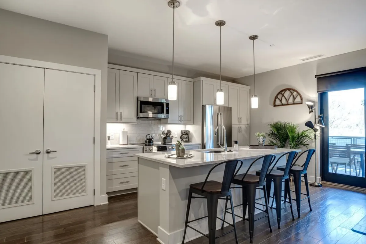 The image showcases a bright, contemporary kitchen with an inviting island counter, sleek bar stools, and modern appliances. Sunlight streams through large windows, hinting at city views and filling the space with warmth. The setting feels perfect for cooking, sharing meals, or enjoying a relaxing coffee in a stylish, urban environment.