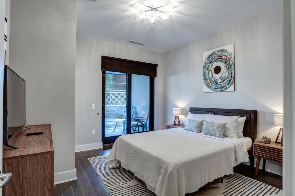 A serene queen bedroom featuring a plush bed with soft bedding, nightstands with contemporary lighting, a window letting in natural light, elegant artwork on the wall, and a television for relaxing evenings. The overall mood is cozy and tranquil, perfect for restful nights.