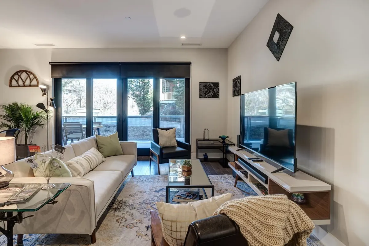 This image showcases a cozy, modern living area featuring a comfortable sofa, coffee table, television, and expansive windows that allow sunlight to pour in. The decor is contemporary, and the setup invites relaxation for movie nights or quiet moments with a cup of coffee.