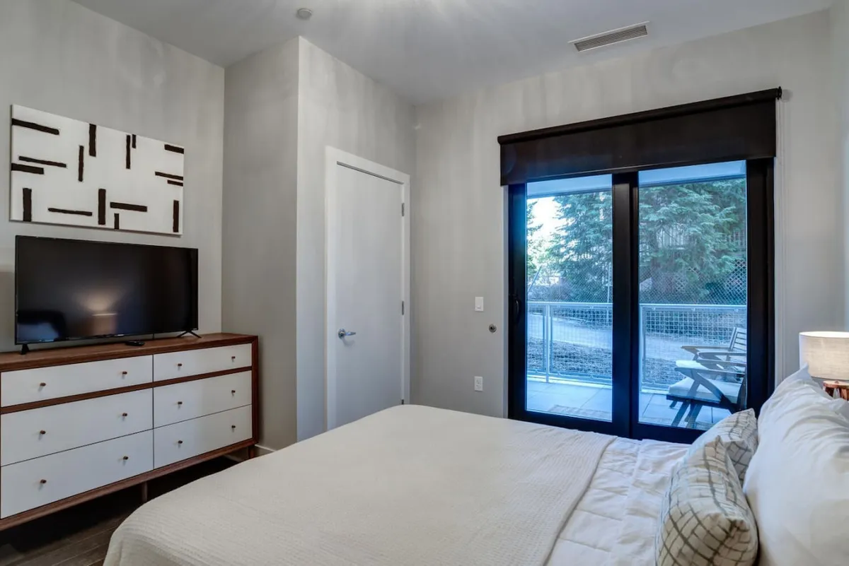 A tranquil bedroom scene featuring a comfortable bed positioned to enjoy views through a sliding glass door, with a dresser and television completing the relaxed ambiance. The space is bathed in natural light, suggesting an inviting transition to the outside world.