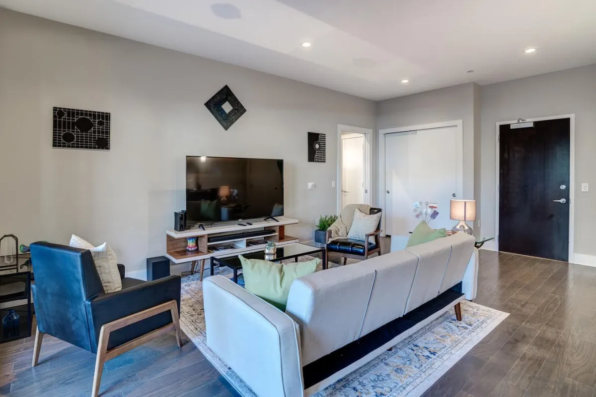 A comfortable, modern living room with a large sofa, a television mounted on the wall, a coffee table, and eye-catching artwork. The space is arranged for relaxation and social gatherings, inviting guests to unwind after exploring downtown.