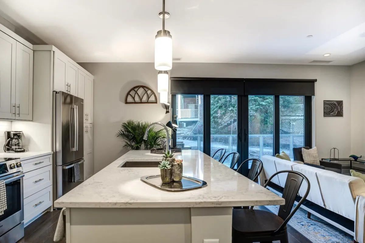 This image highlights a contemporary kitchen with a central island, large windows that fill the space with natural light, modern stainless steel appliances, and touches of greenery from decorative plants. The scene captures people cooking together and hints at social gatherings, making the kitchen feel lively and welcoming.