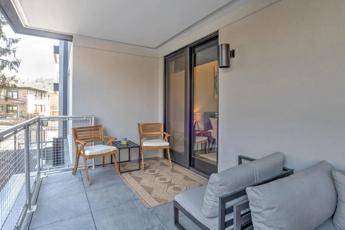 A cozy exterior balcony furnished with comfortable seating, a textured rug, and a small table, all set against a peaceful city backdrop visible through glass doors. The warm, tranquil mood is ideal for morning coffee, reading, or intimate gatherings, with soft lighting suggesting early morning or sunset.